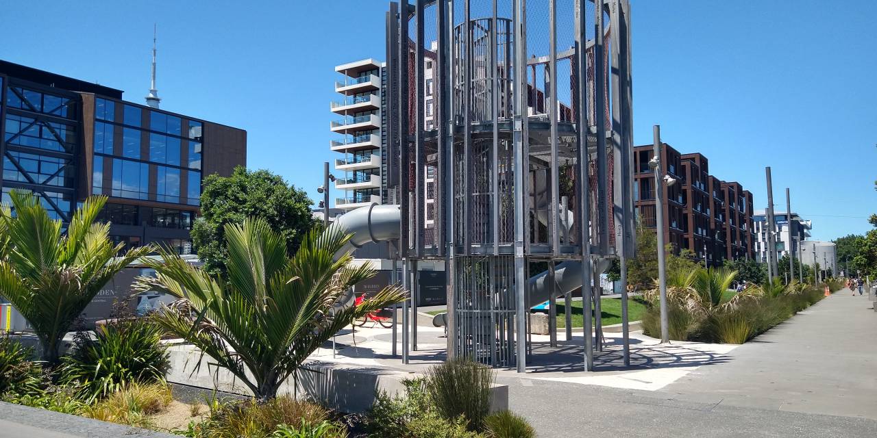 Daldy Street linear park play area: an industrial steel structure with slide surrounded by planting with blue sky in the background. In Wynyard Quarter, Auckland.