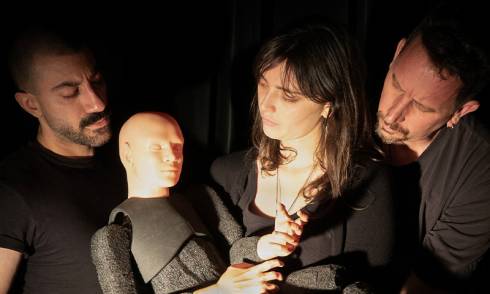 Two men and a woman are low lit against a black background and are peering down at a small, faceless puppet that the woman is holding