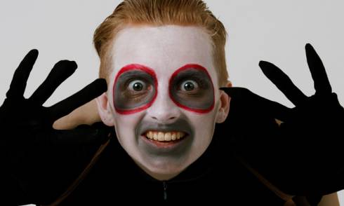 A man with white facepaint and black circles around his eyes bares his teeth at the camera with raised hands