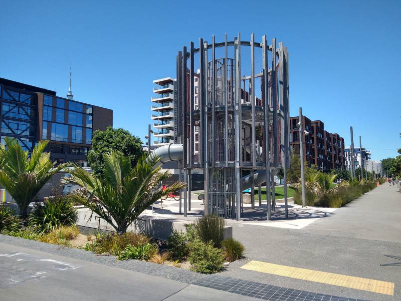 Daldy Street linear park play area: an industrial steel structure with slide surrounded by planting with blue sky in the background. In Wynyard Quarter, Auckland.